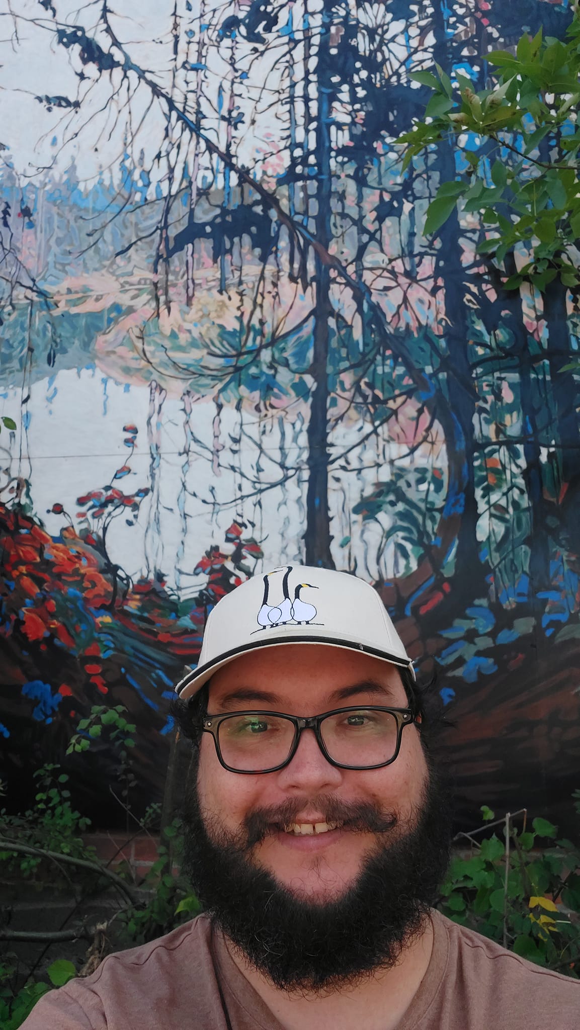 A man with short hair and bushy facial hair stands in front of an outdoor mural of Tom Tompson's painting "Northern River"